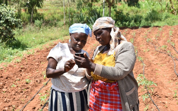 Dos mujeres de pie en un campo. La mujer situada a la derecha sostiene un teléfono móvil con la mano izquierda. Las dos miran el teléfono.