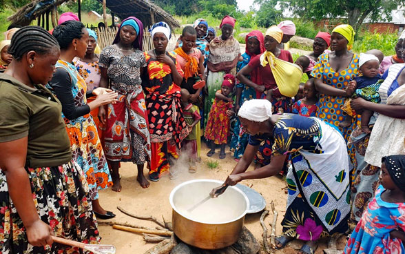 Many women surround a cauldron over a fire in Tanzania. In the centre, a cook stirs the water and the grains to be cooked.