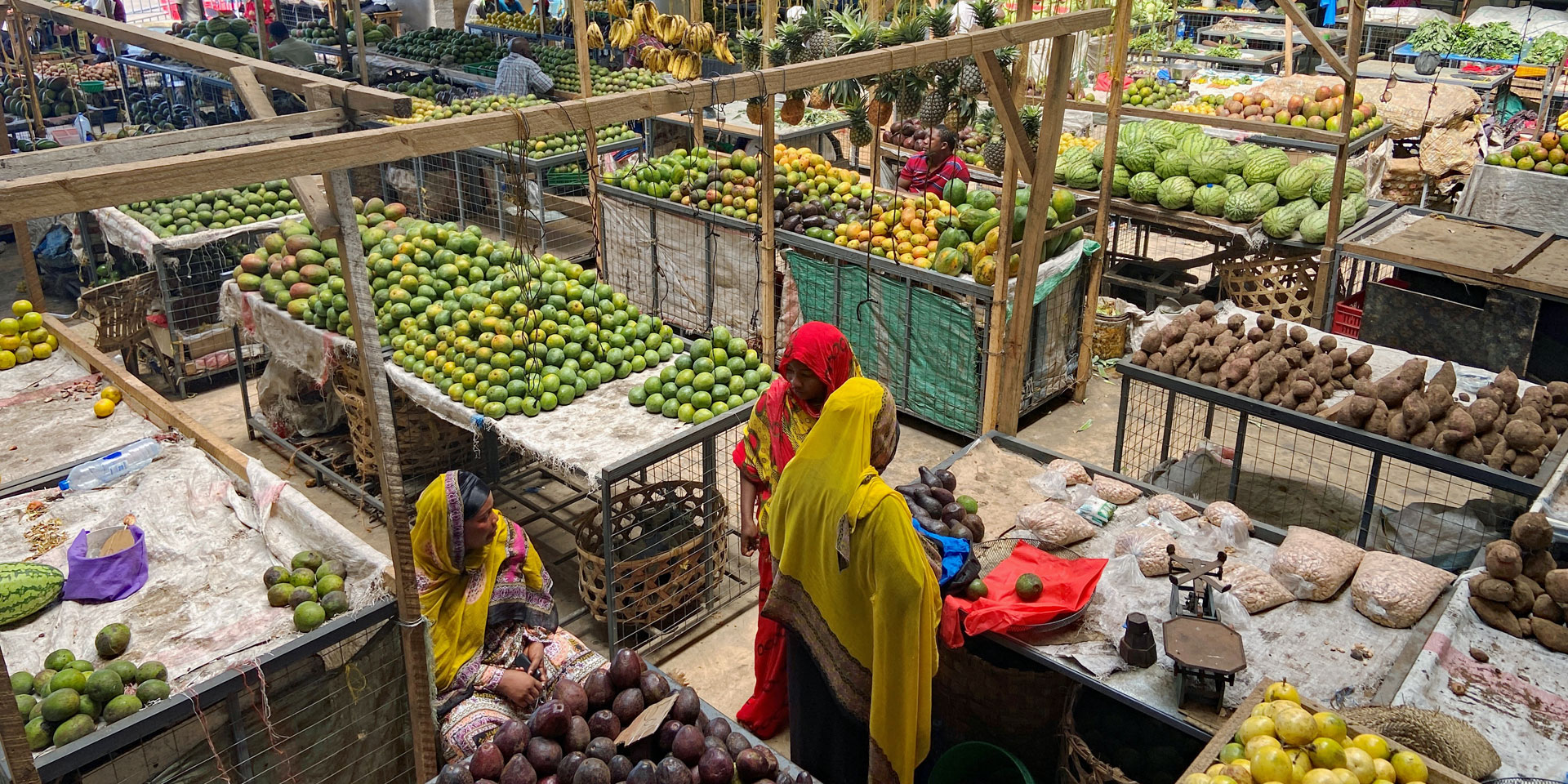 Deux femmes voilées de rouge et de jaune se tiennent devant leur stand de fruits. Des rayons remplis de fruits et légumes les entourent.