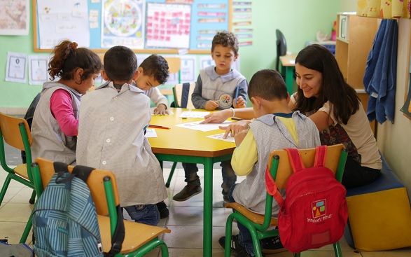 Foto dell’interno di una classe con un’insegnante in piedi mentre mostra un libro a un gruppo di bambine e bambini.