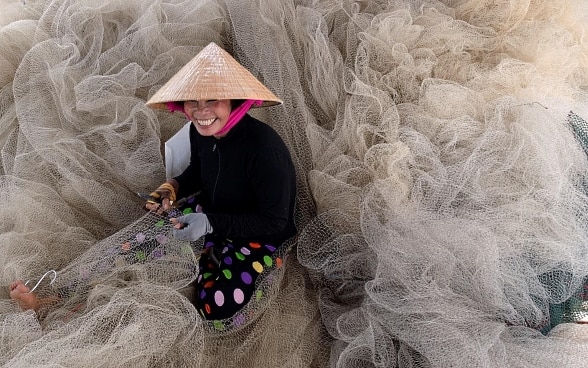 Une jeune femme est assise au milieu de filets de pêche. Elle sourit et porte un chapeau. Elle tient un outil dans une main et de l’autre, elle répare un filet de pêche.