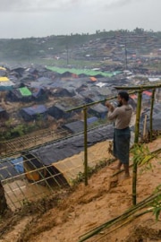 A man stands on the edge of a refugee camp in Cox’s Bazar. He is building a shelter for his family out of lengths of wooden pole.