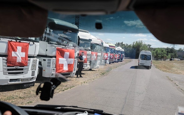 Zehn Lastwagen mit Schweizer Flaggen stehen aufgereiht auf einem Feld in der Ukraine. 